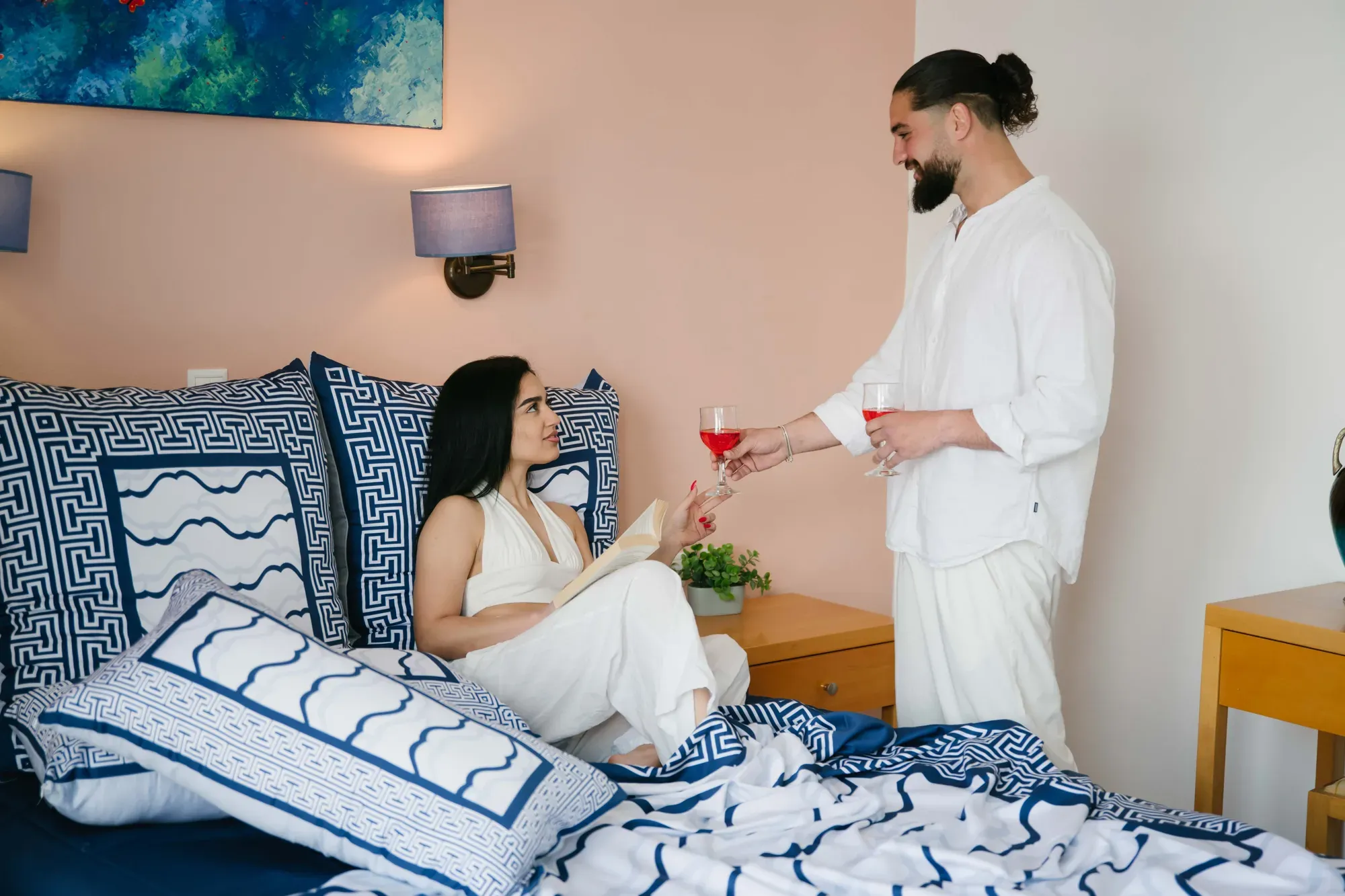 A man brings a glass to a woman sitting up in a bed made in Cycladic Serenity