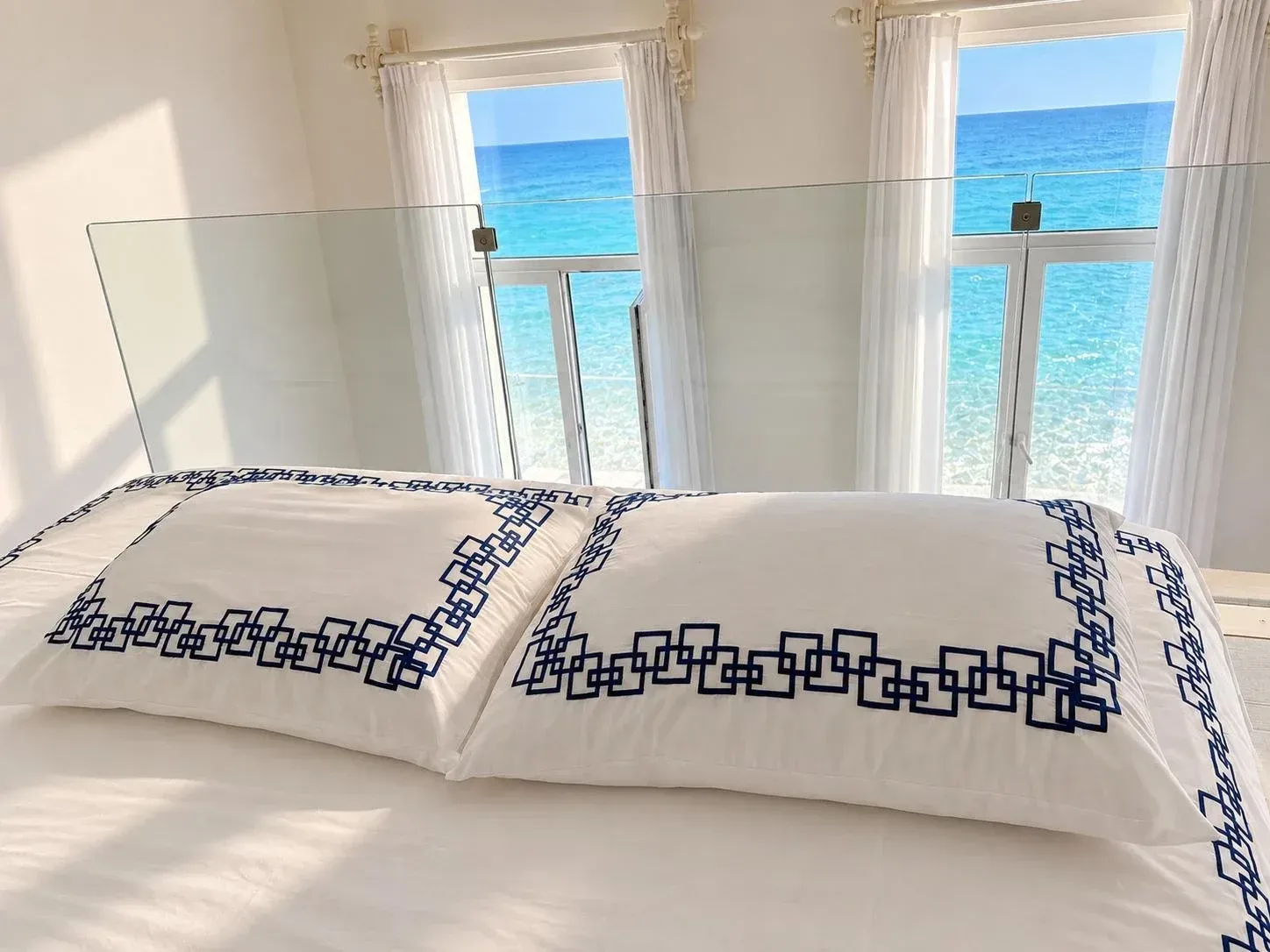Two Desmos pillowcases on a white bed, the sea bright through the windows